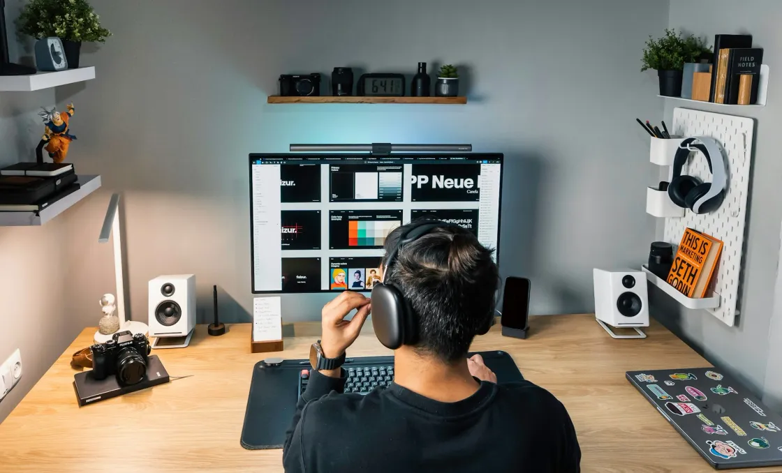 Designer wearing headphones reviews color palettes and typography on a widescreen monitor in a tidy home studio workspace.