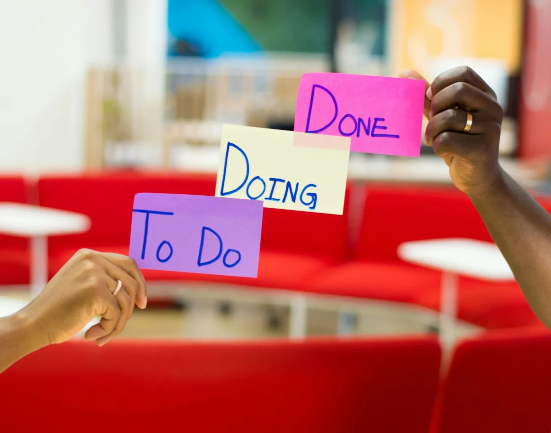 Two hands hold sticky notes reading “To Do,” “Doing,” and “Done,” stacked Kanban-style to show a project workflow in progress.