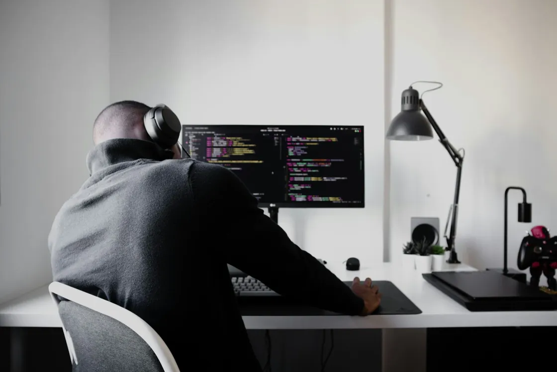 Back-view photo of a person wearing over-ear headphones and coding on a wide monitor at a tidy white desk, with a lamp, plants, and game controller in view.