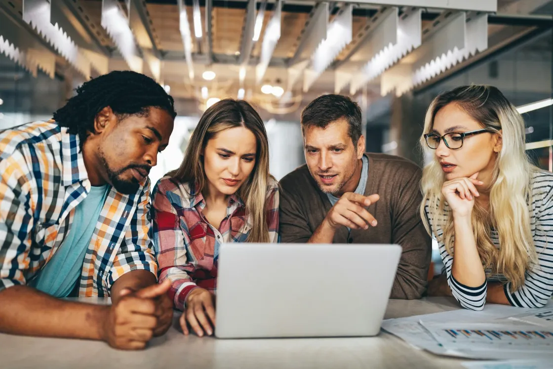 Four coworkers gathered around a laptop, reviewing a shared document and charts in a modern workspace; illustrates asynchronous communication and team collaboration.