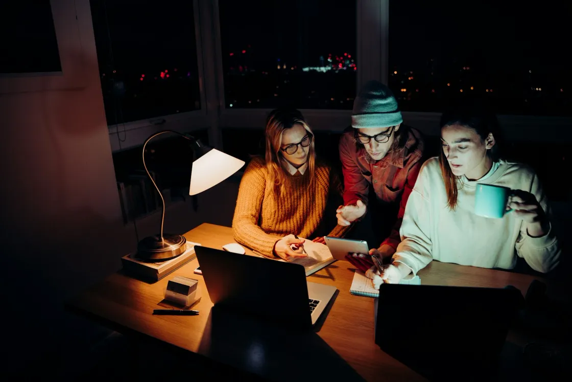 Three professionals work at night with laptops and a tablet under a desk lamp, illustrating remote team collaboration across time zones in Singapore, London and New York.