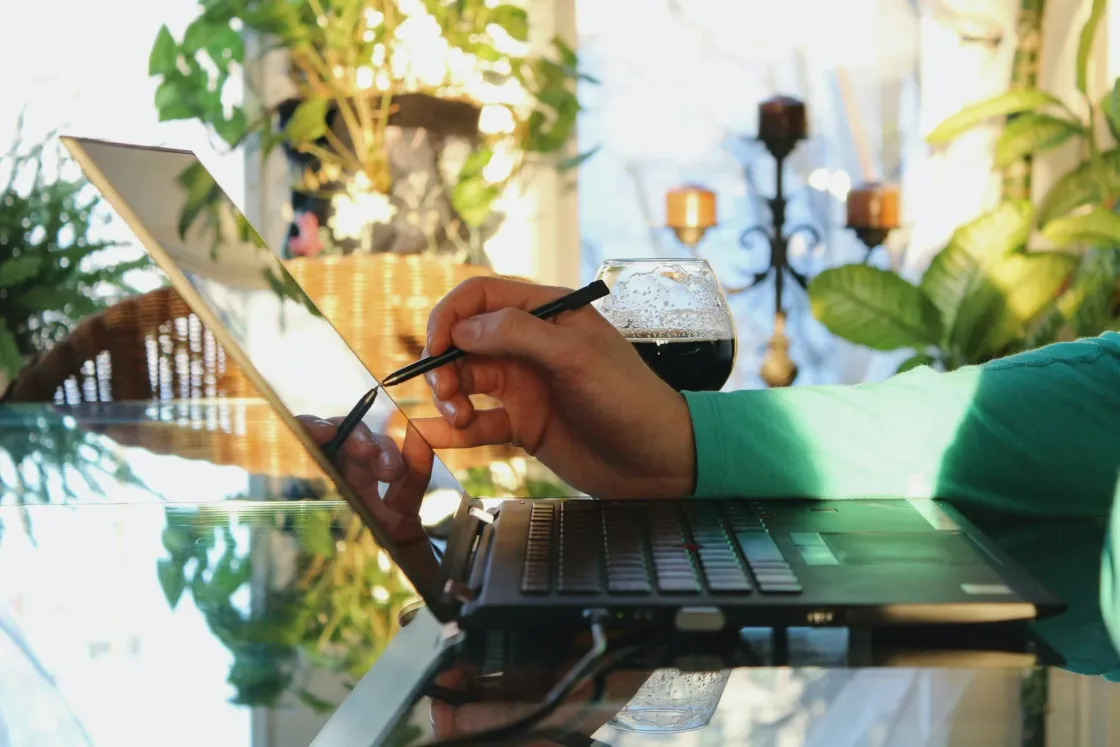 Designer uses a stylus on a touchscreen laptop at a sunlit home desk, preparing a Figma case study with a drink nearby and plants in the background.
