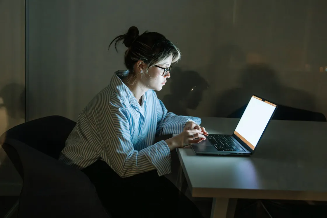 Remote-first culture scene: a focused woman in glasses works on a laptop at night in a quiet office, screen glow highlighting async collaboration and deep work.