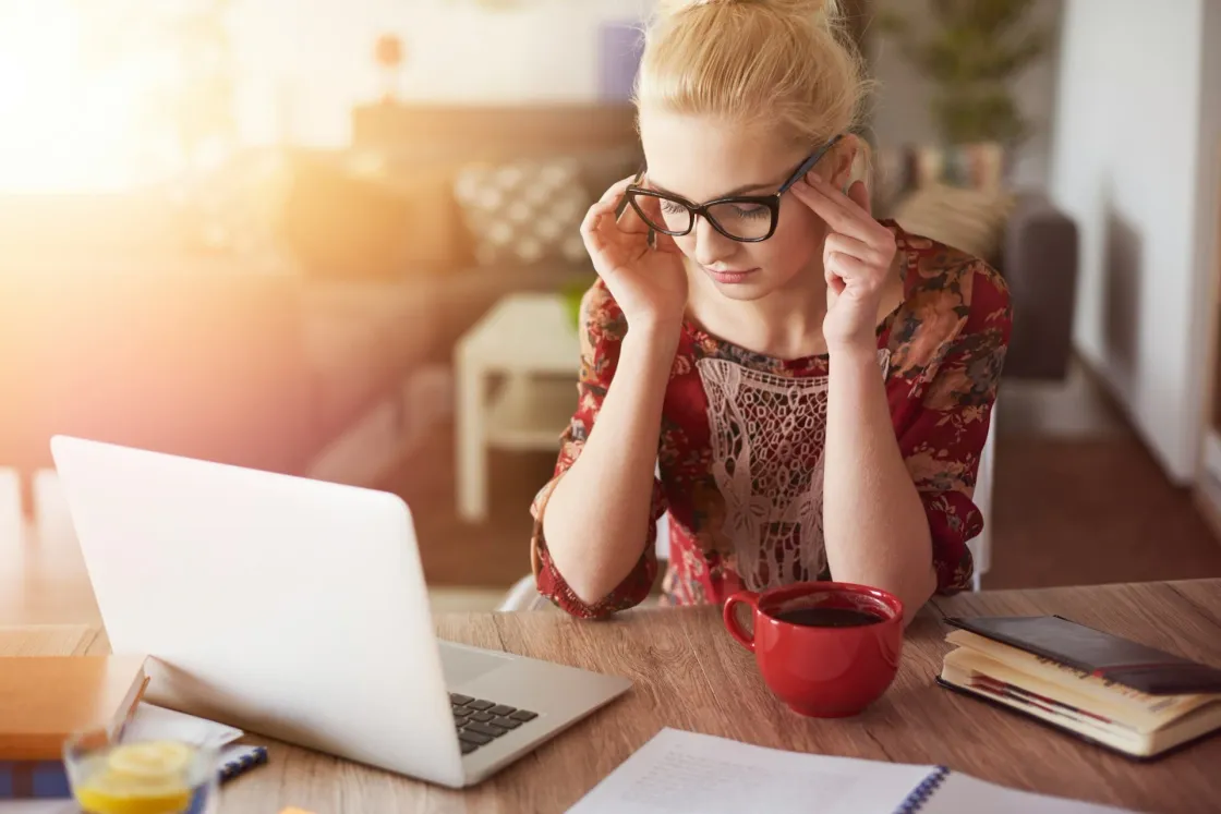 Person working on a laptop at a home desk, adjusting glasses, with a red coffee mug and notebook nearby.