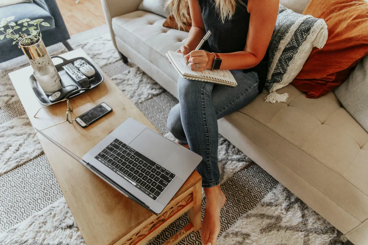 Person writing notes on a sofa beside an open laptop and phone on a coffee table in a calm home workspace.