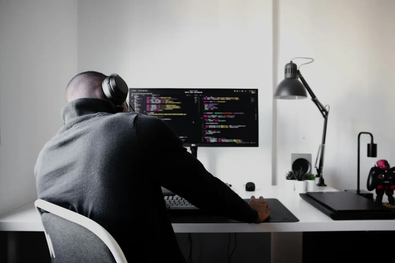 Back-view photo of a person wearing over-ear headphones and coding on a wide monitor at a tidy white desk, with a lamp, plants, and game controller in view.