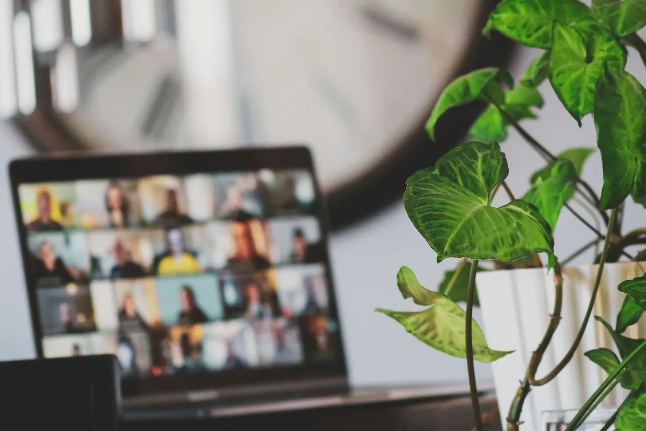 Laptop with a video call grid blurred in the background and a green plant in the foreground, illustrating a hybrid team standup.