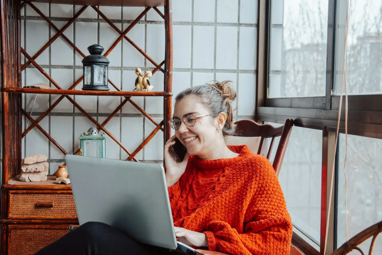 Person working from home on a laptop while talking on the phone, sitting by a window.